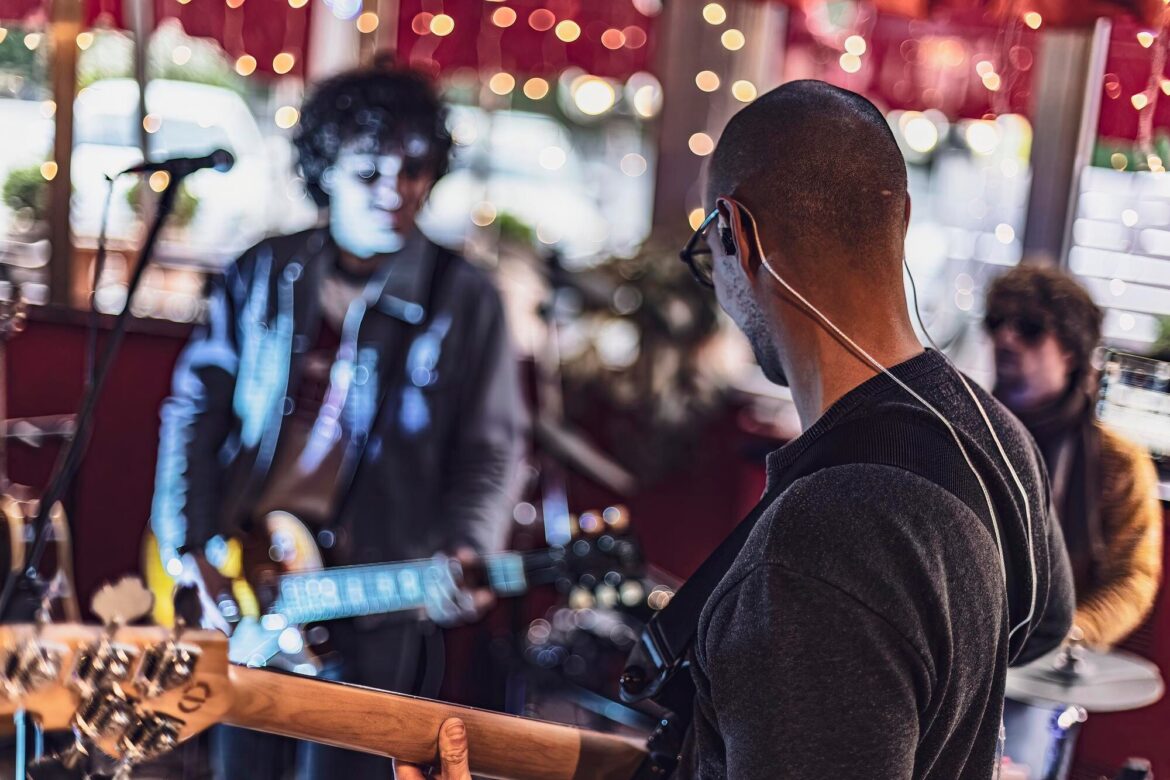 Bassist and guitarist performing with a drummer in a small venue lit by string lights.